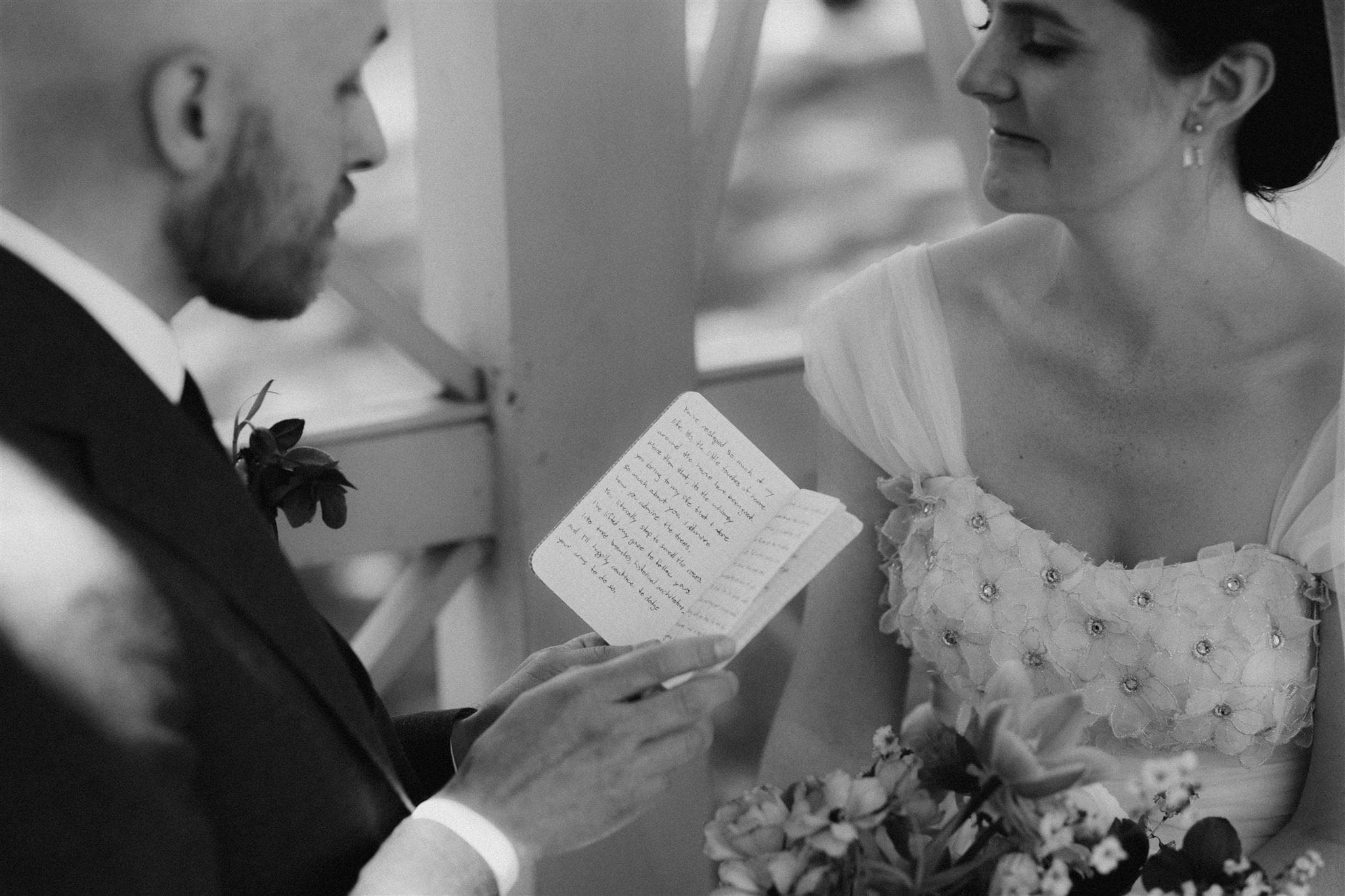 Black and white image of a married couple sitting close together on their Philadelphia wedding day while groom reads handwritten vows from a small notebook