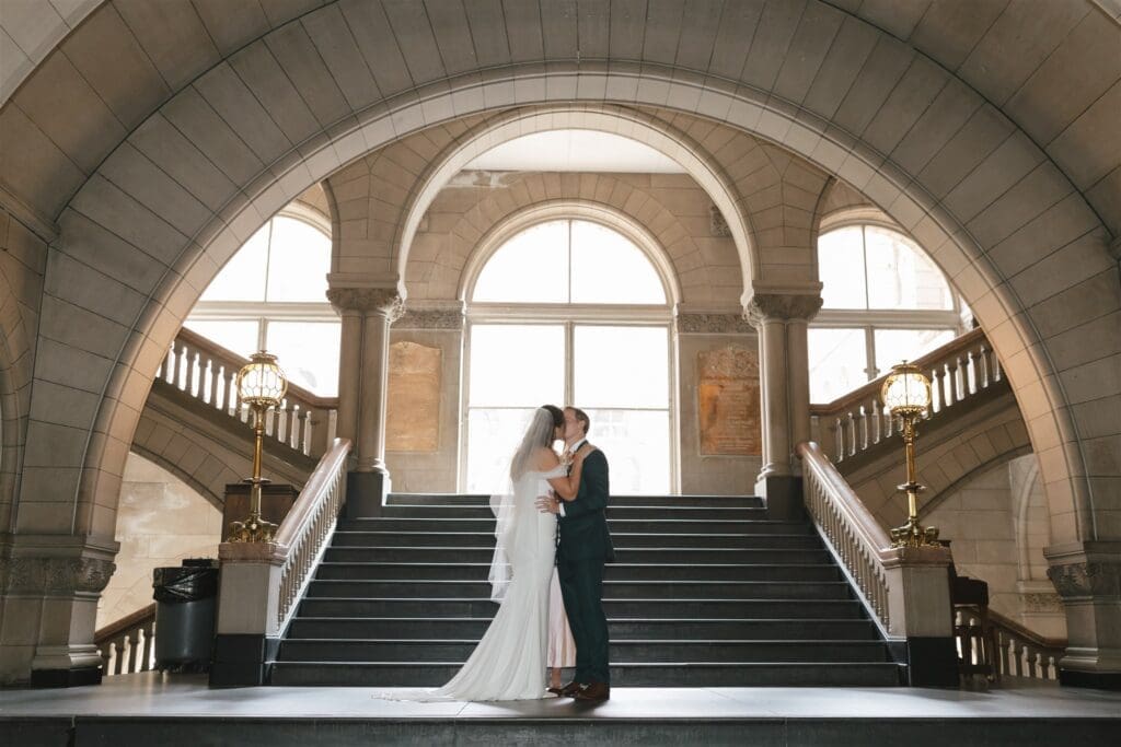 Bride and groom kissing at the base of the Grand Staircase inside Allegheny County Courthouse in Pittsburgh, Pennsylvania — wide architectural wedding portrait framed by a sweeping stone arch, brass lanterns, and dual staircases rising on either side