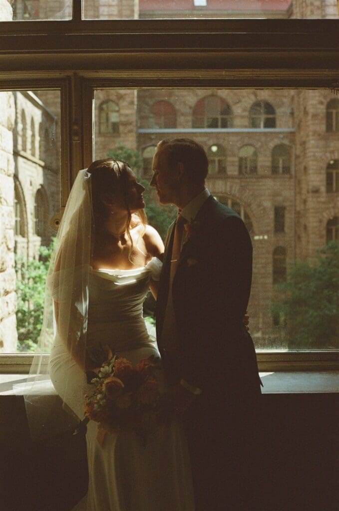 Moody film portrait of a bride and groom sitting on a window sill kissing with a historic stone building in the backdrop