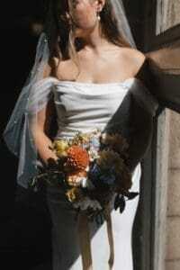 Close-up bridal portrait of bride holding a garden wedding bouquet of terracotta dahlias, blue delphinium, peach roses, and white daisies against her off-shoulder crepe gown — warm directional light outside the ornate iron doors of Allegheny County Courthouse in Pittsburgh