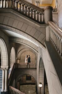 Bride and groom kissing on the mezzanine landing of the Grand Staircase at Allegheny County Courthouse in Pittsburgh, Pennsylvania — small figures framed within layered stone arches, a signature architectural wedding portrait by Pennsylvania wedding photographer Shiloh Leath
