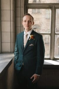 Groom portrait in forest green suit with terracotta and peach boutonnière, photographed by a tall window inside Allegheny County Courthouse in Pittsburgh, Pennsylvania — clean, natural light wedding portrait