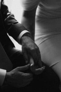 Black and white film-inspired close-up of groom gently holding bride's hand against her off-shoulder wedding gown — quiet, intimate wedding detail shot inside Allegheny County Courthouse in Pittsburgh, Pennsylvania
