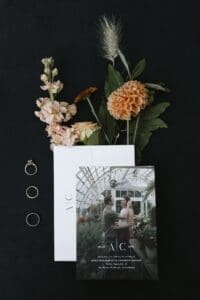 Wedding detail flatlay for an intimate Pittsburgh wedding at Bar Marco — invitation suite with engagement photo, three gold wedding rings, peach dahlia, pink stock flowers, and pampas grass on a dark background