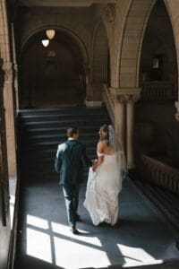 Bride and groom walking away from camera through a sun-drenched corridor inside Allegheny County Courthouse in Pittsburgh, Pennsylvania — candid intimate wedding moment with dramatic window light geometry on the stone floor