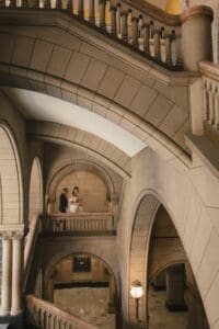 Bride and groom standing together on the mezzanine landing of the Grand Staircase inside Allegheny County Courthouse in Pittsburgh, Pennsylvania — wide architectural wedding portrait showing the full sweep of stone arches and ornate lanterns