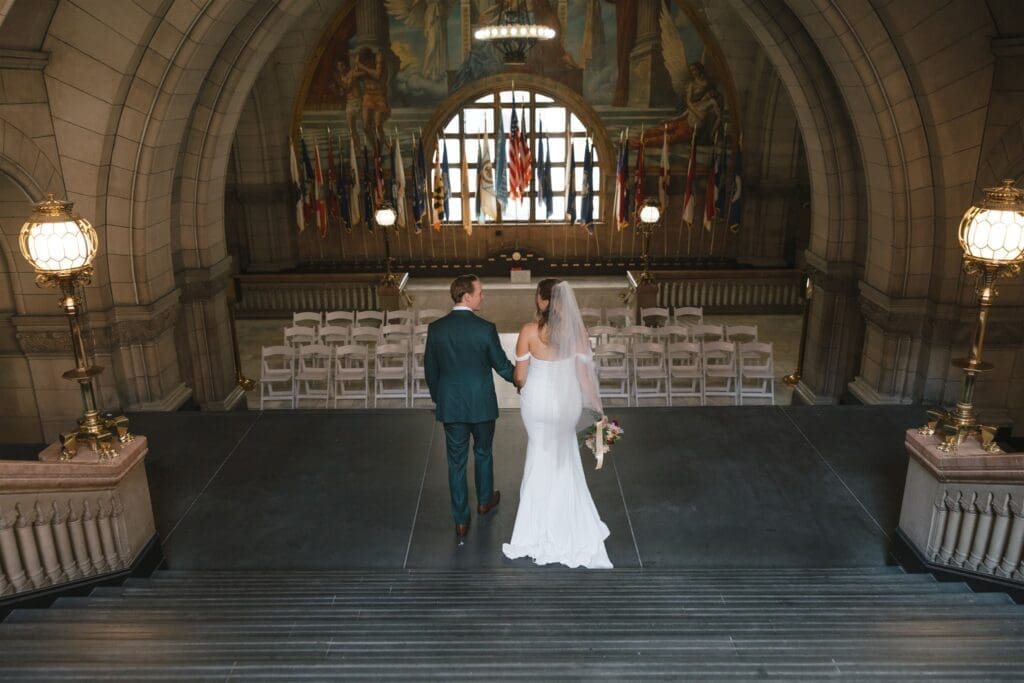 Bride and groom walking hand in hand toward the ceremony room inside Allegheny County Courthouse in Pittsburgh — state flags flanking an arched window, photographed from above in a documentary, film-inspired style