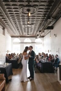Bride and groom sharing their first kiss during the first dance at Union Hall at Bar Marco in Pittsburgh's Strip District — wide reception shot showing guests seated at long tables beneath the tin ceiling and arched windows