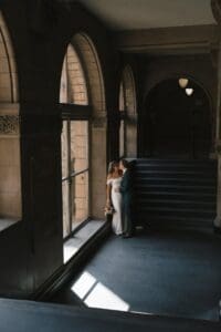Bride and groom sharing a quiet kiss beside a tall Gothic window inside Allegheny County Courthouse in Pittsburgh — moody, light-pooling intimate wedding portrait by hybrid film and digital photographer Shiloh Leath