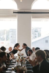 Candid wedding reception moment at Union Hall at Bar Marco in Pittsburgh — guests laughing and connecting at a long banquet table with warm window light flooding the intimate second-floor gallery space