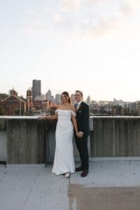 Bride and groom standing together on a rooftop overlooking the Pittsburgh skyline at golden hour, bride in off-shoulder crepe gown and groom in forest green suit — intimate wedding portrait by a Pennsylvania wedding photographer
