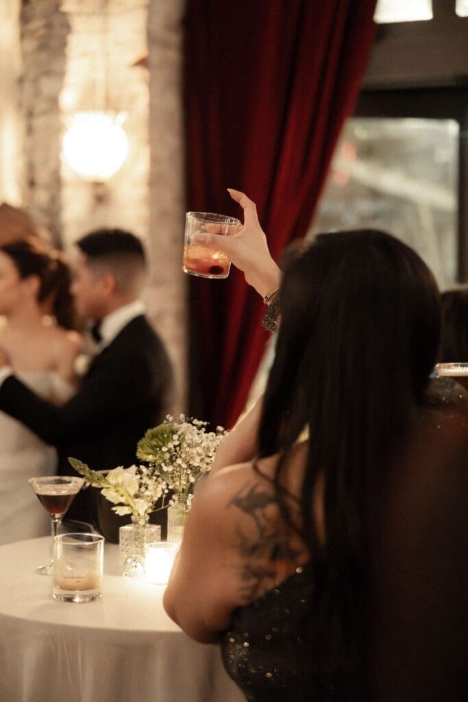 A guest raising her cocktail as the newlyweds embrace during the wedding reception