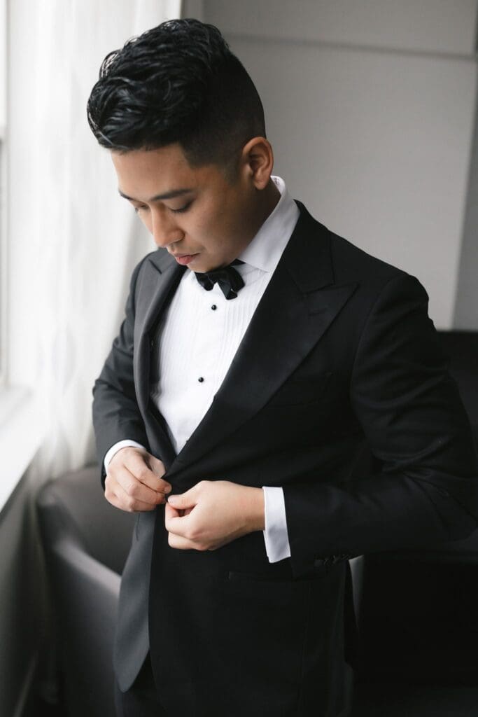 Groom buttoning his tuxedo jacket as he finishes getting ready
