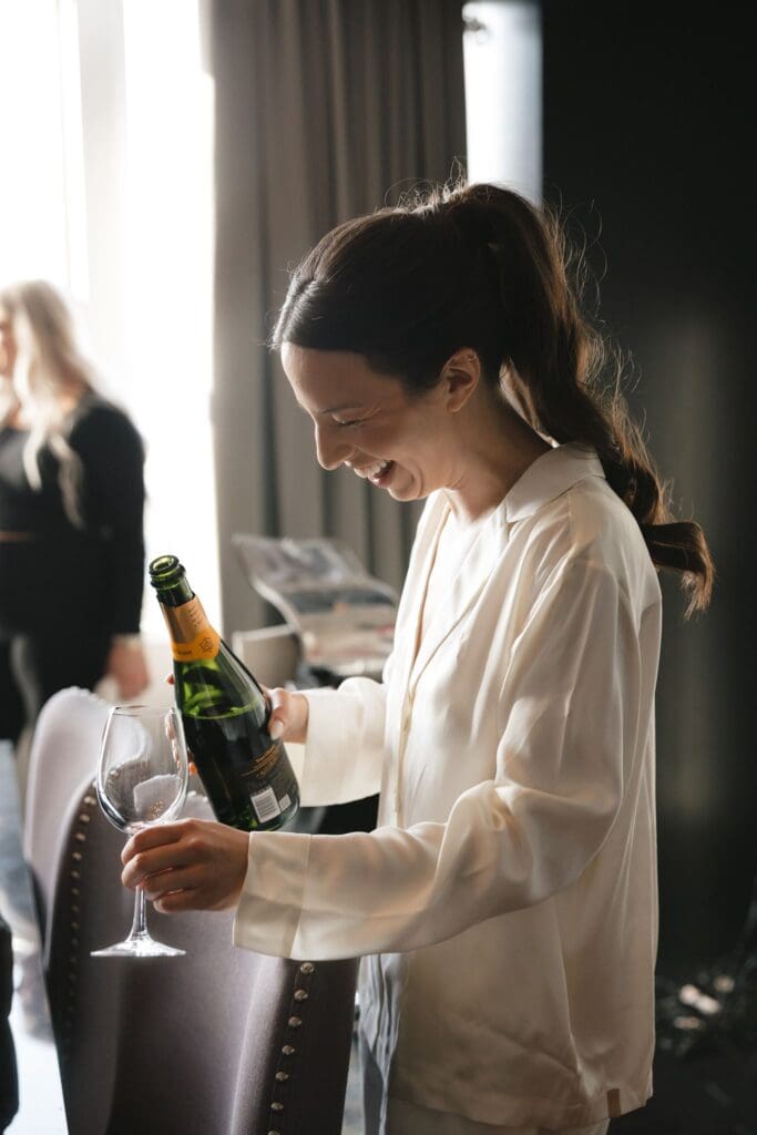 Bride laughing as she pours champagne while getting ready