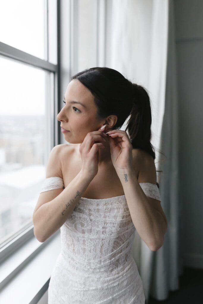 Bride fastening an earring by the window in an off-the-shoulder lace wedding dress