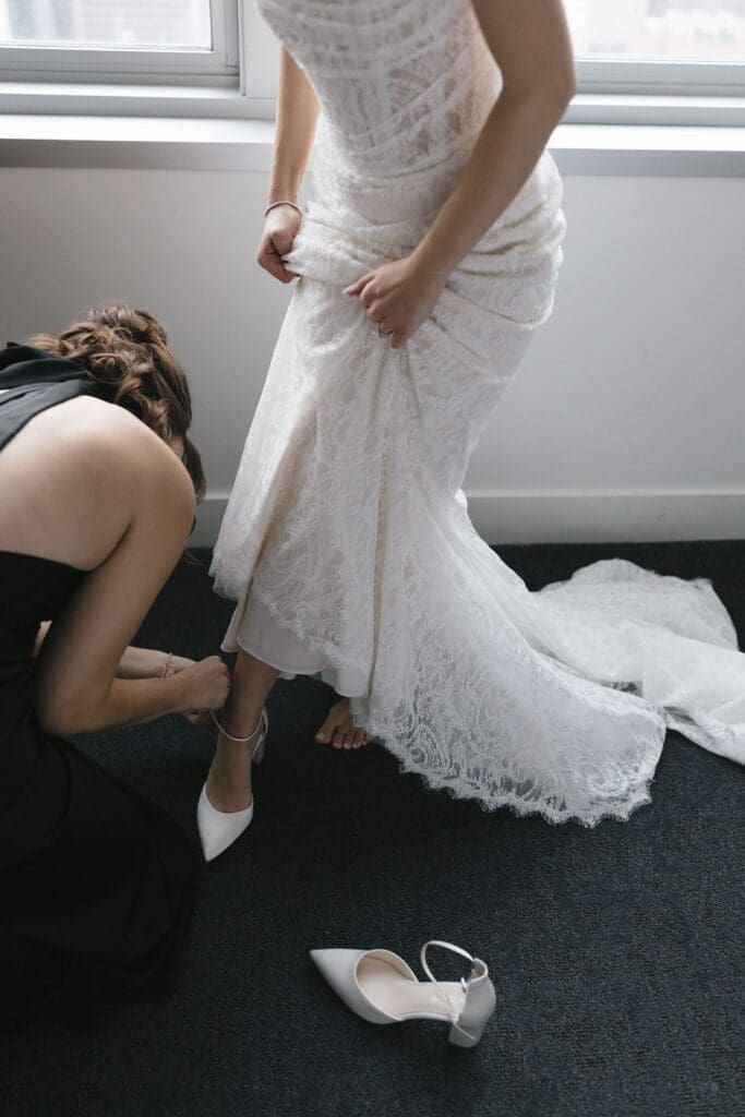 A bridesmaid buckling the bride's shoe, her lace wedding dress train pooled across the floor