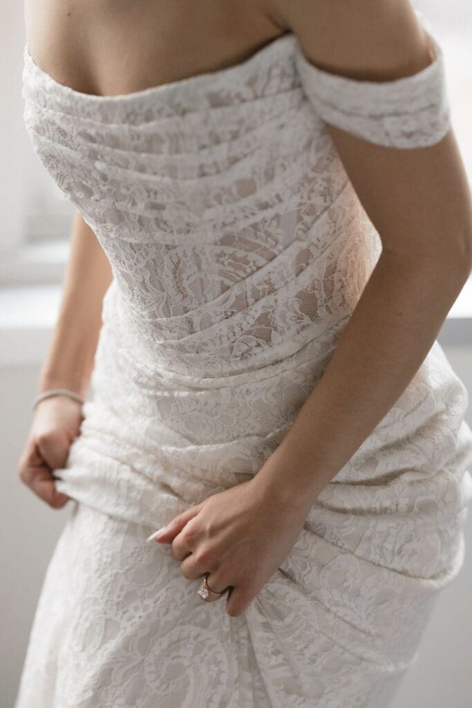 Detail of an off-the-shoulder lace wedding dress bodice as the bride smooths the fabric
