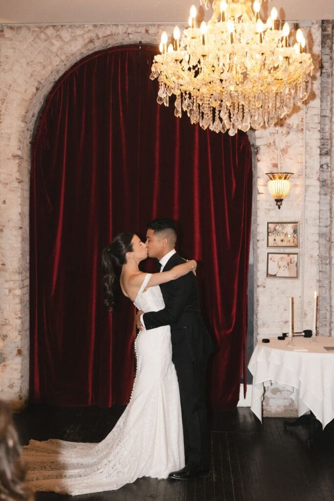 Bride and groom's first kiss beneath a crystal chandelier and red velvet curtain at the Divine Lorraine