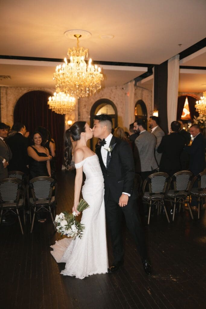 Newlyweds kissing beneath a crystal chandelier as guests look on after the ceremony