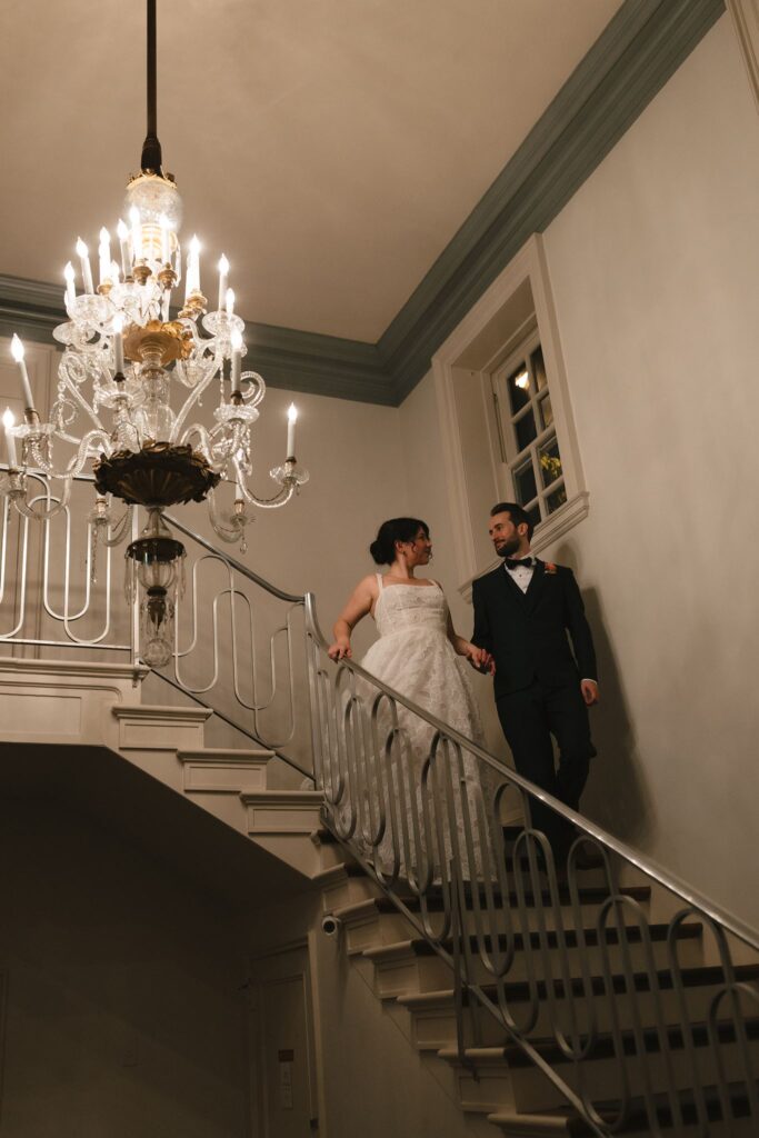 Bride and groom walk down a staircase next to an ornate vintage chandelier at the Inn at Fox Briar Farm
