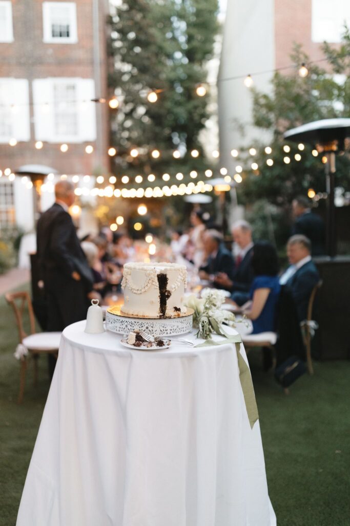 Single tier white wedding cake with one sliver removed sits on a small cocktail table with wedding guests lounging at a table in the background