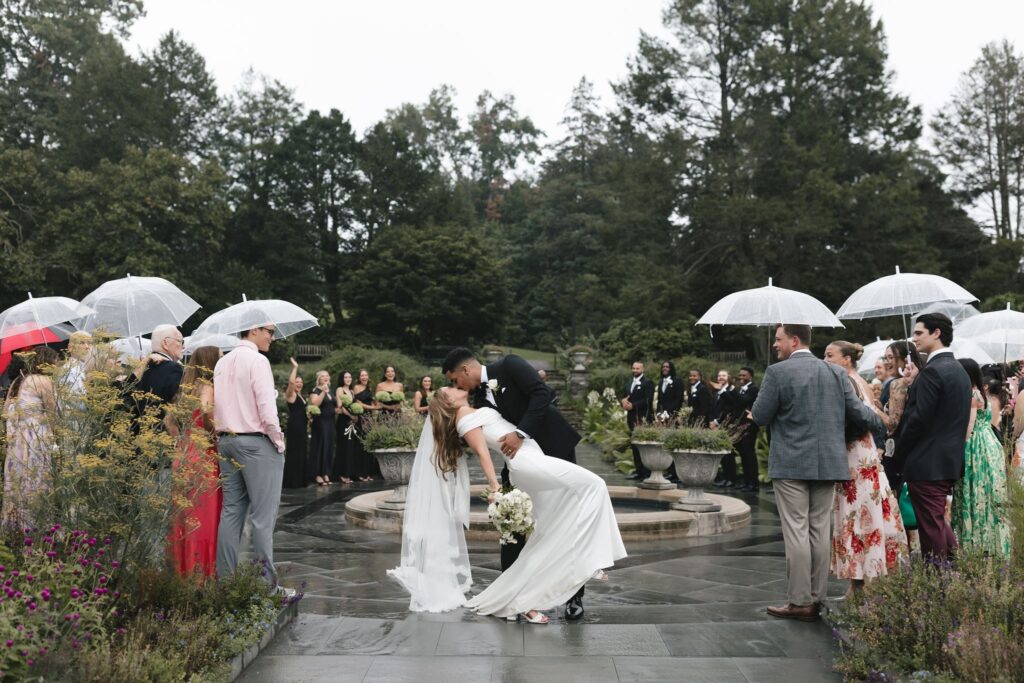 Groom dips bride for a kiss while wedding guests holding clear umbrellas look on during a rainy ceremony at Morris Arboretum