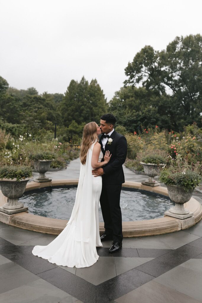 Bride and groom kiss in front of a fountain surrounded by flowers in urn planters at Morris Arboretum
