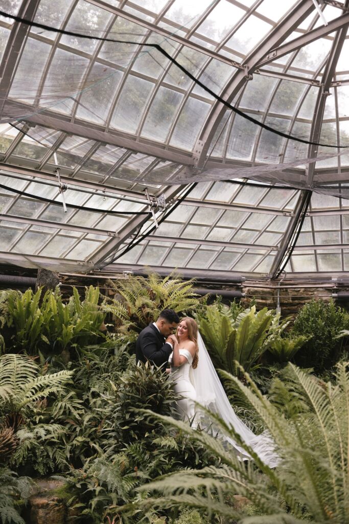 Bride and groom stand in a greenhouse at the historic Morris Arboretum surrounded by vibrant green ferns