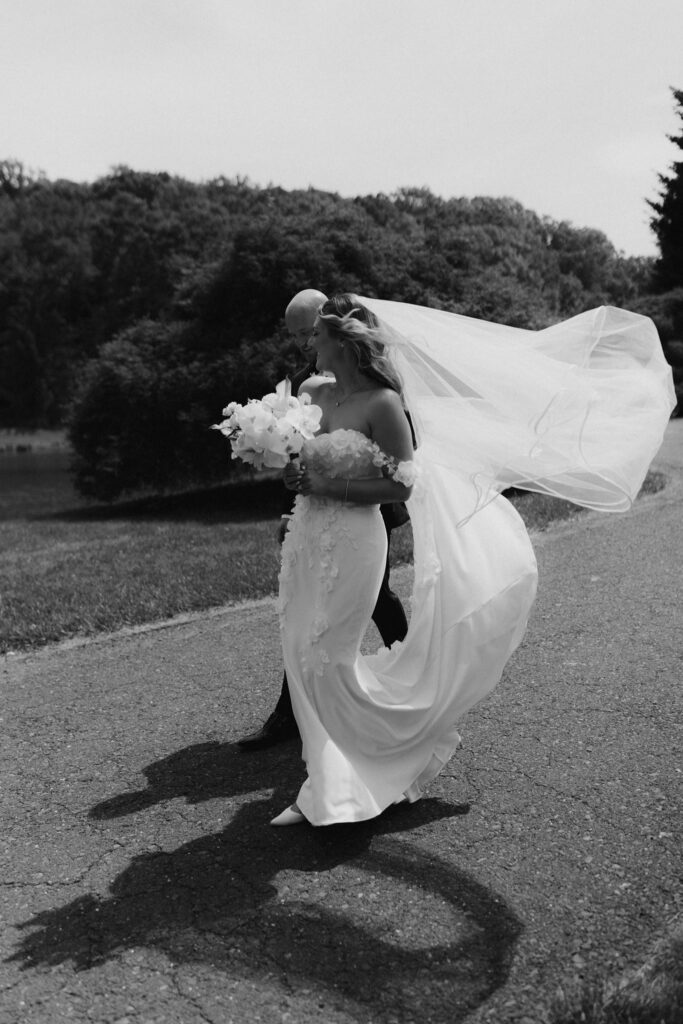 Black and white portrait of a bride and groom walking down a driveway with bride's veil flowing behind them