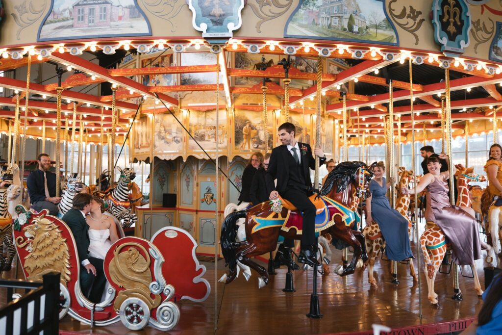 Wedding guests riding on a carousel watch the bride and groom kiss at a unique wedding venue, the Carousel at Pottstown in Pennsylvania