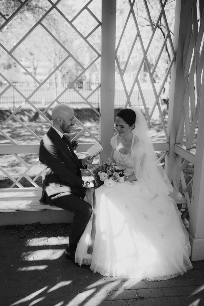 Groom reads vows from a small book to his smiling bride while sitting on a bench under a pergola in the 18th Century Garden in Philadelphia's Old City