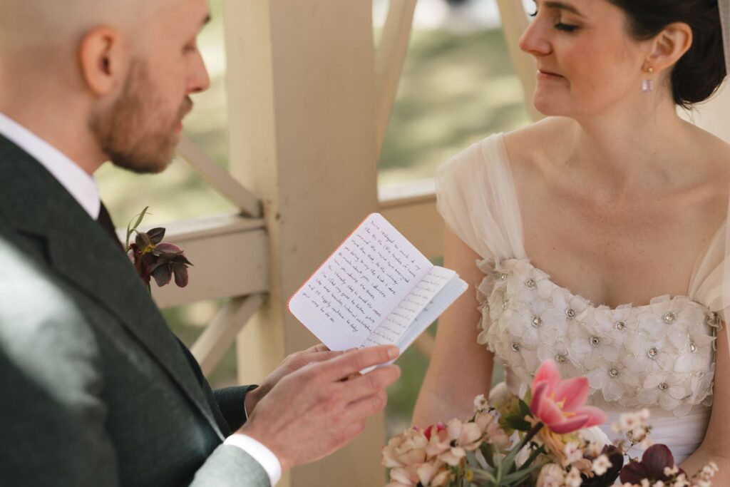 Groom reads vows from a small book to his bride under a pergola in the 18th Century Garden in Philadelphia's Old City