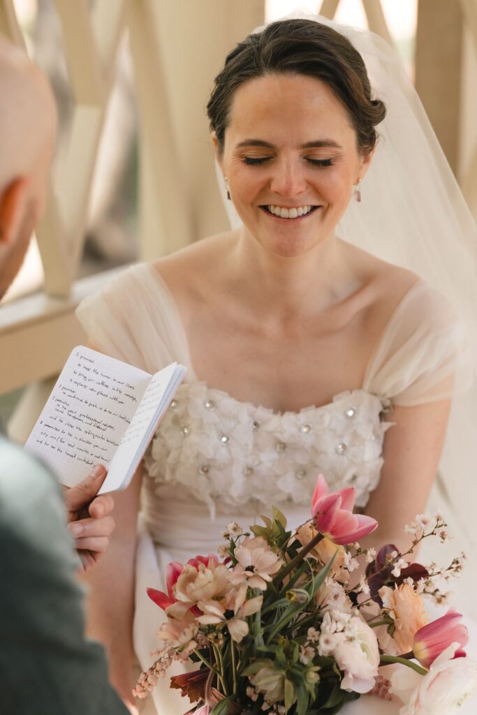 Groom reads vows from a small book to his smiling bride under a pergola in the 18th Century Garden in Philadelphia's Old City