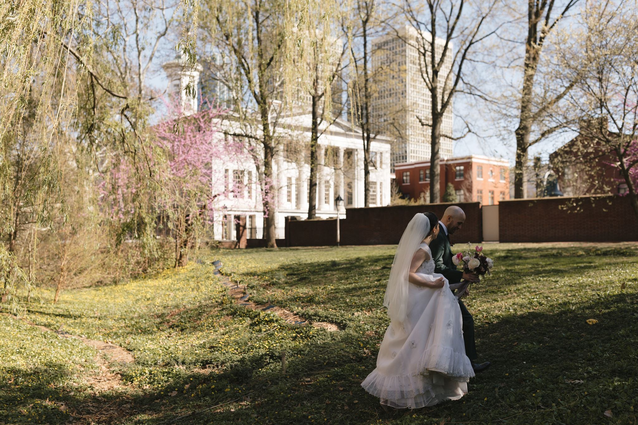 Bride and groom walk under willow trees in a park in Philadelphia with historic buildings in the background | Shiloh Leath Photography
