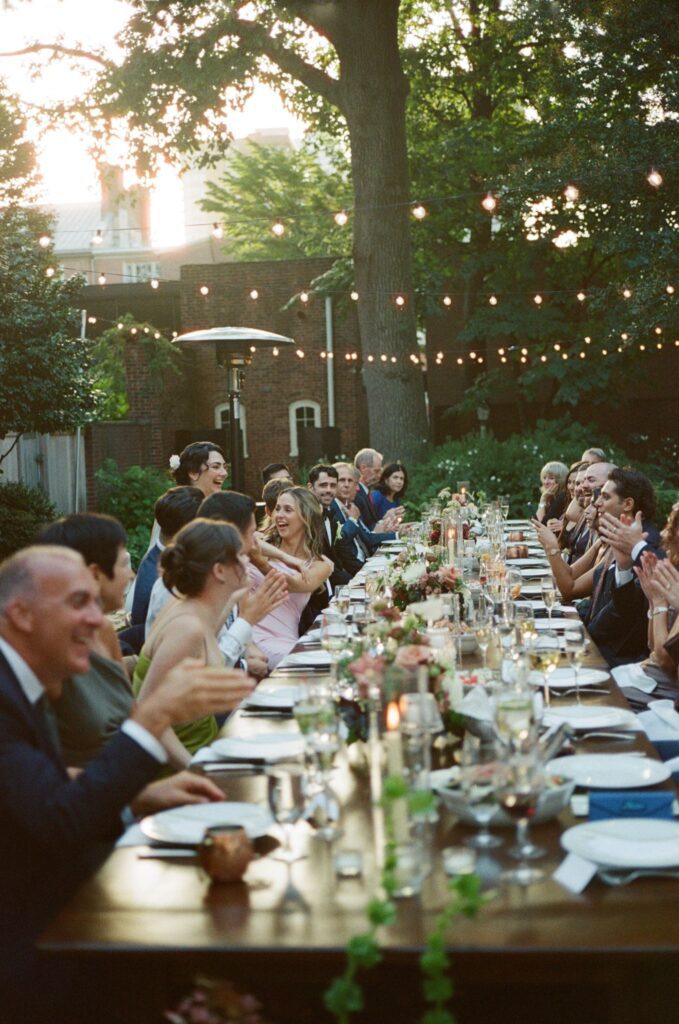 Guests gather and laugh around a long banquet table under market lights during a late afternoon backyard wedding at the Powel House in Philadelphia