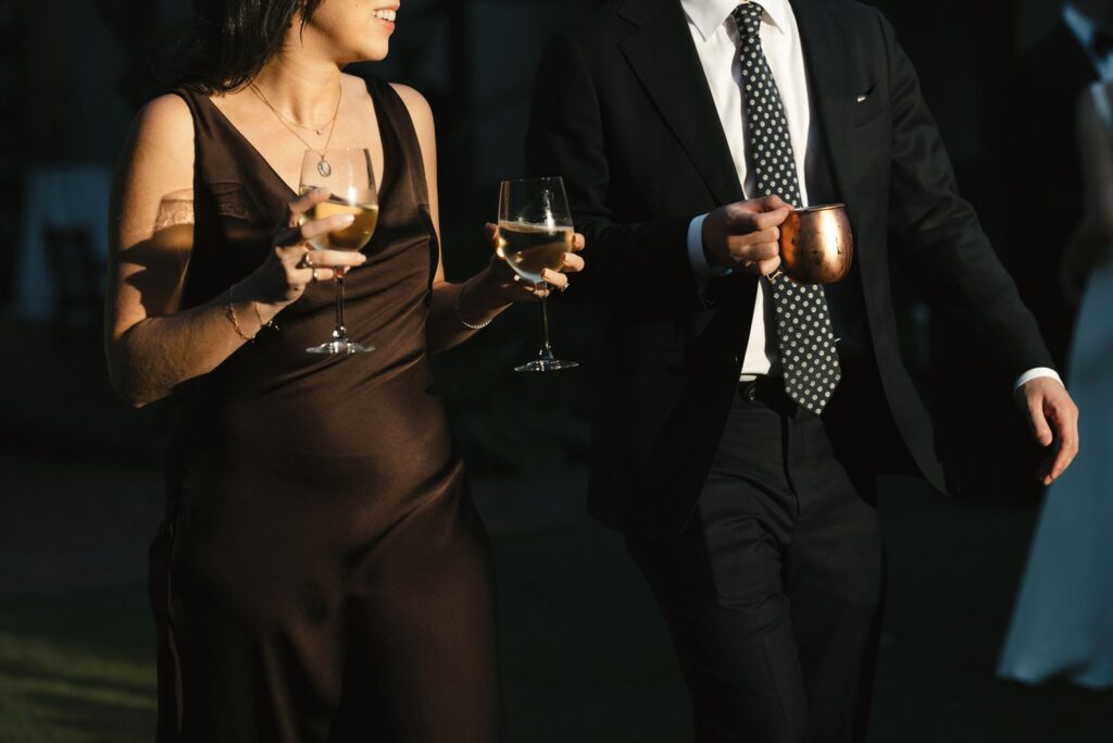 Two wedding guests walk together while carrying glasses of wine and an Old Fashioned cocktail from the bar