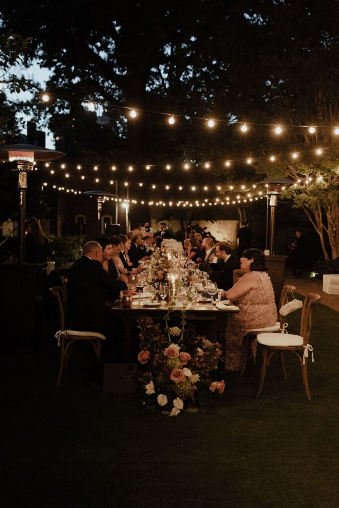 Wedding guests sit at a long banquet table at night under cafe lights and outdoor heaters