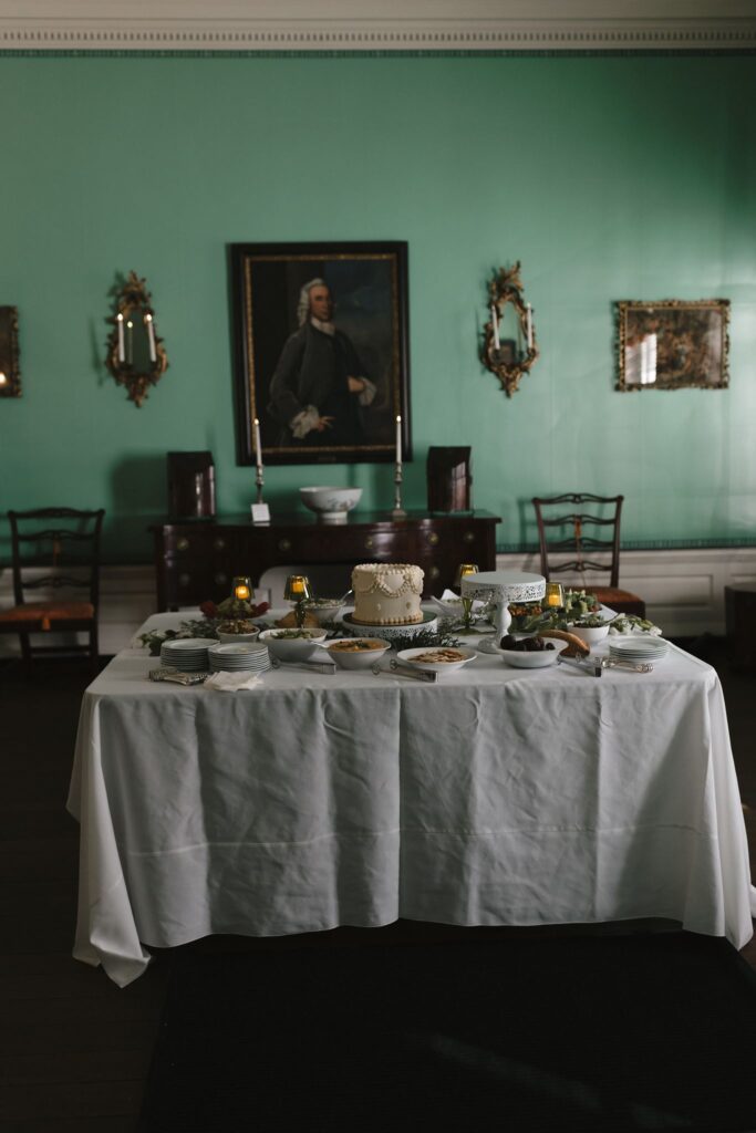 Table set with hors d'oeuvres, small plates, and a single tier buttercream wedding cake sits in a mint green room with 18th century artwork and decor at the Powel House